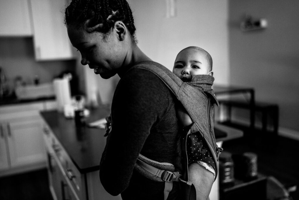 A mom walks around her apartment with a baby on her back in a baby carrier during an in home family photography session in Fairfax VA