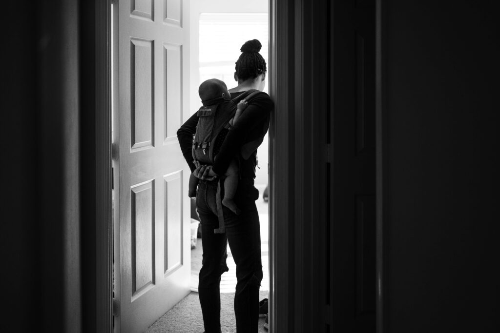 A mom wearing a baby on her back in a baby carrier while looking into her sons bedroom during a documentary family photography session in Fairfax, VA
