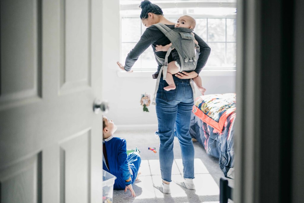 A mom wearing a baby on her back in a baby carrier while in her son's bedroom during a documentary family photography session in Fairfax, VA