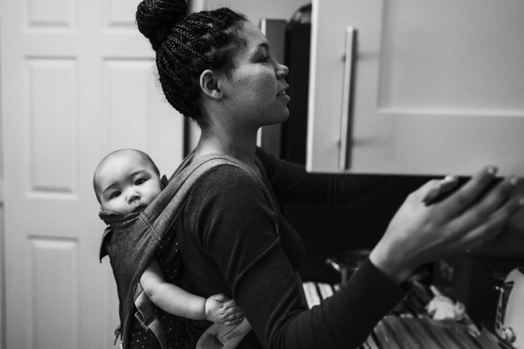 A mom prepares a snack in a kitchen and looks into a kitchen cabinet while wearing her baby on her back during a family photography session