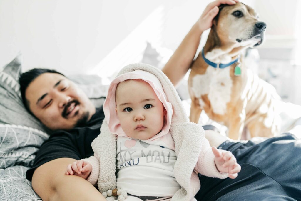 A man pets his dog in bed while he snuggles with his baby during an in-home family photography session