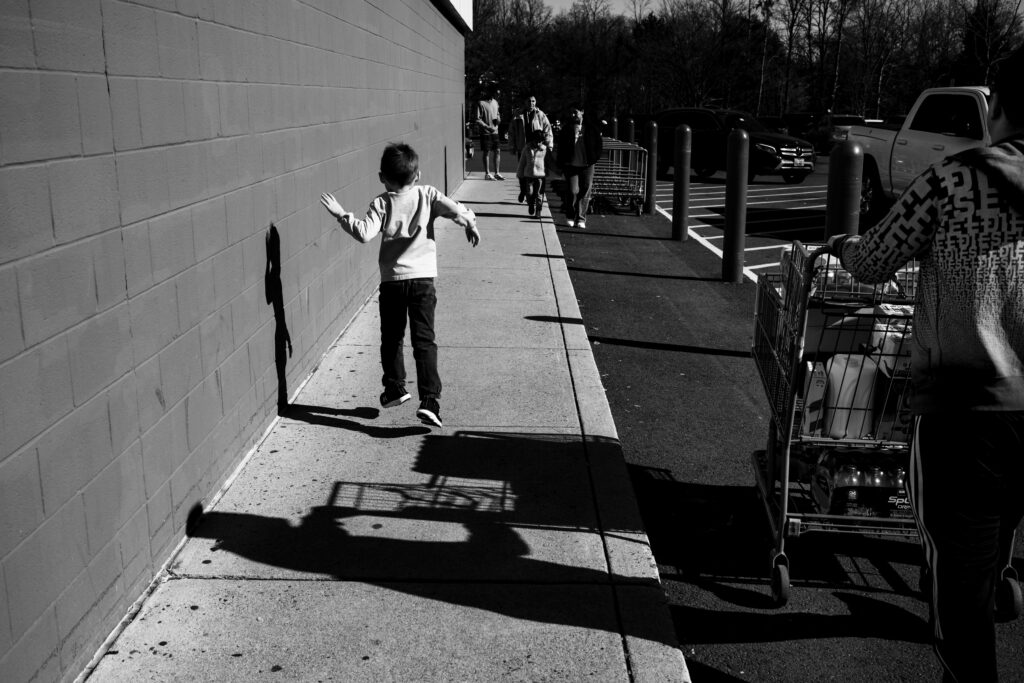 The shadow of a jumping boy outside of COSTCO while his dad pushes the shopping cart in the parking lot A boy sits in a shopping cart while his dad pushes it during a trip to COSTCO during a documentary family photography session by These Days Photography in Fairfax, VA
