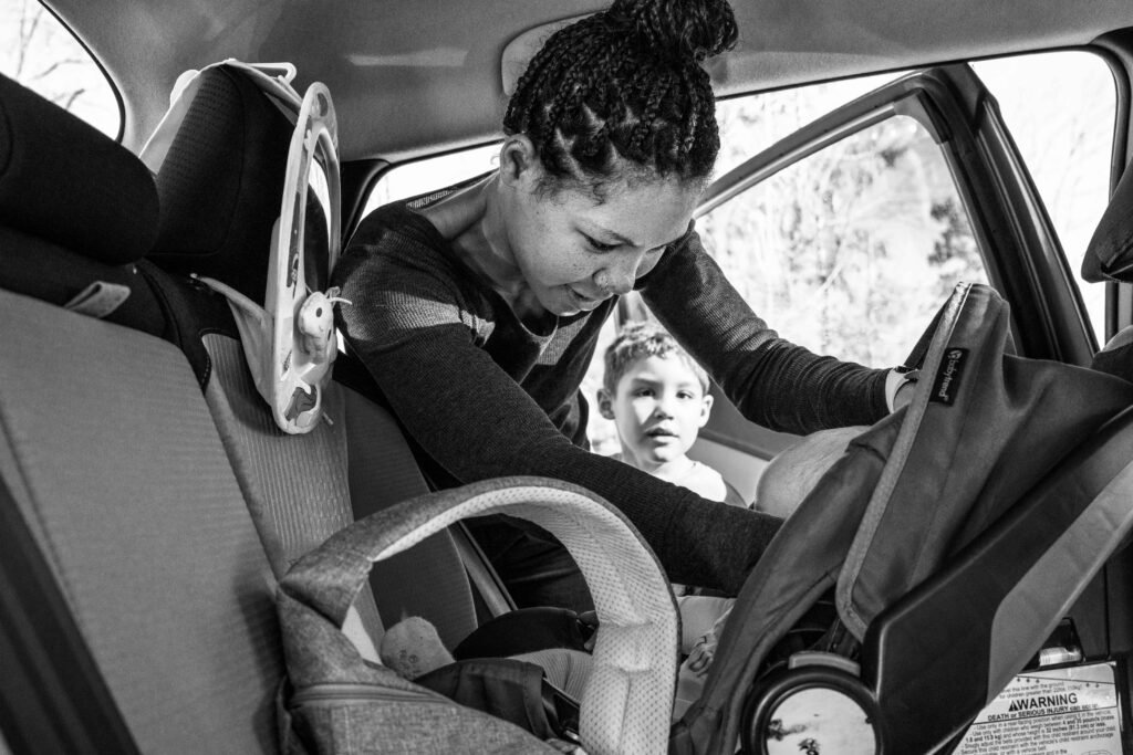 A mom puts her baby into their carseat with her son watching while he stands by the car during a documentary family photography session in Fairfax, Va