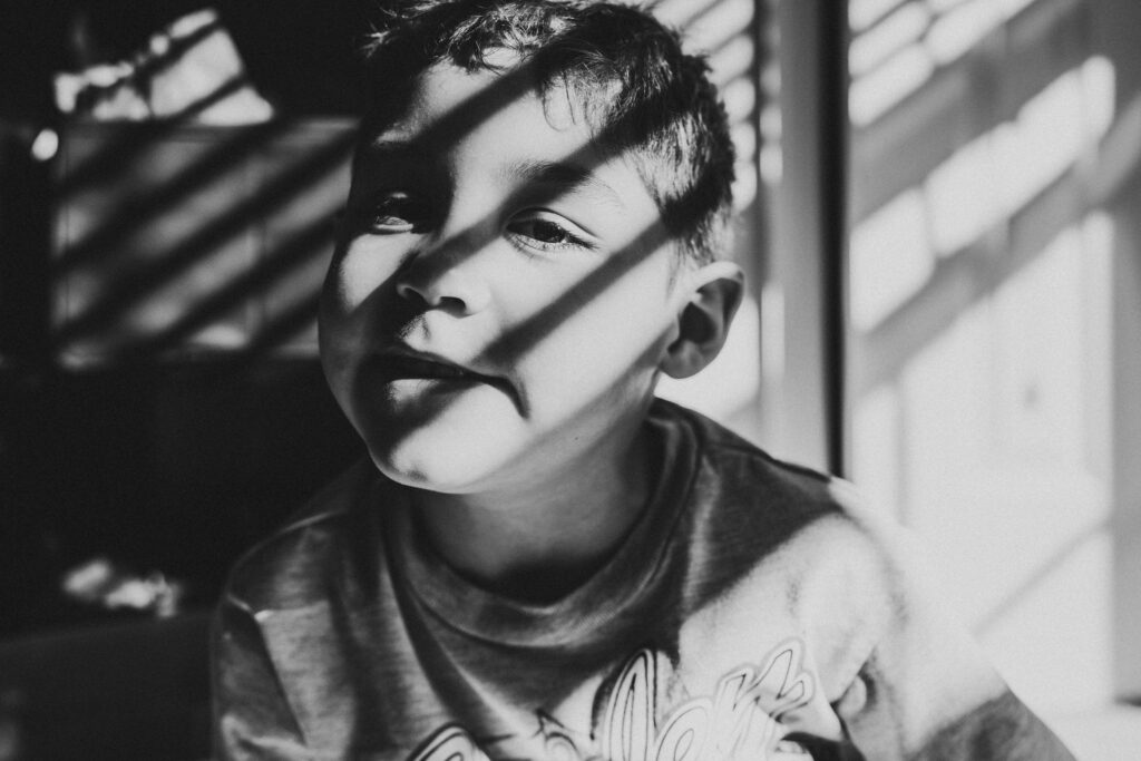 A boy looks at the camera with dark shadows across his face from the sun coming through the blinds during a documentary family photography session by These Days Photography in Fairfax, VA
