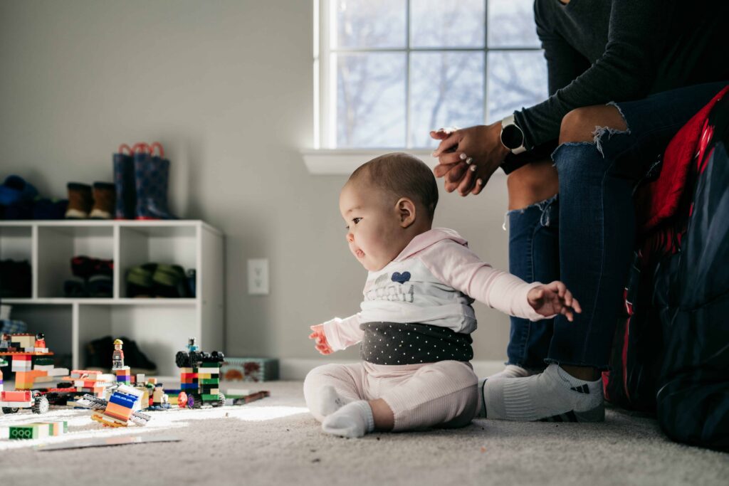 A baby plays with legos while her mother sits on the bed behind her during a documentary family photography session by These Days Photography in Fairfax, VA