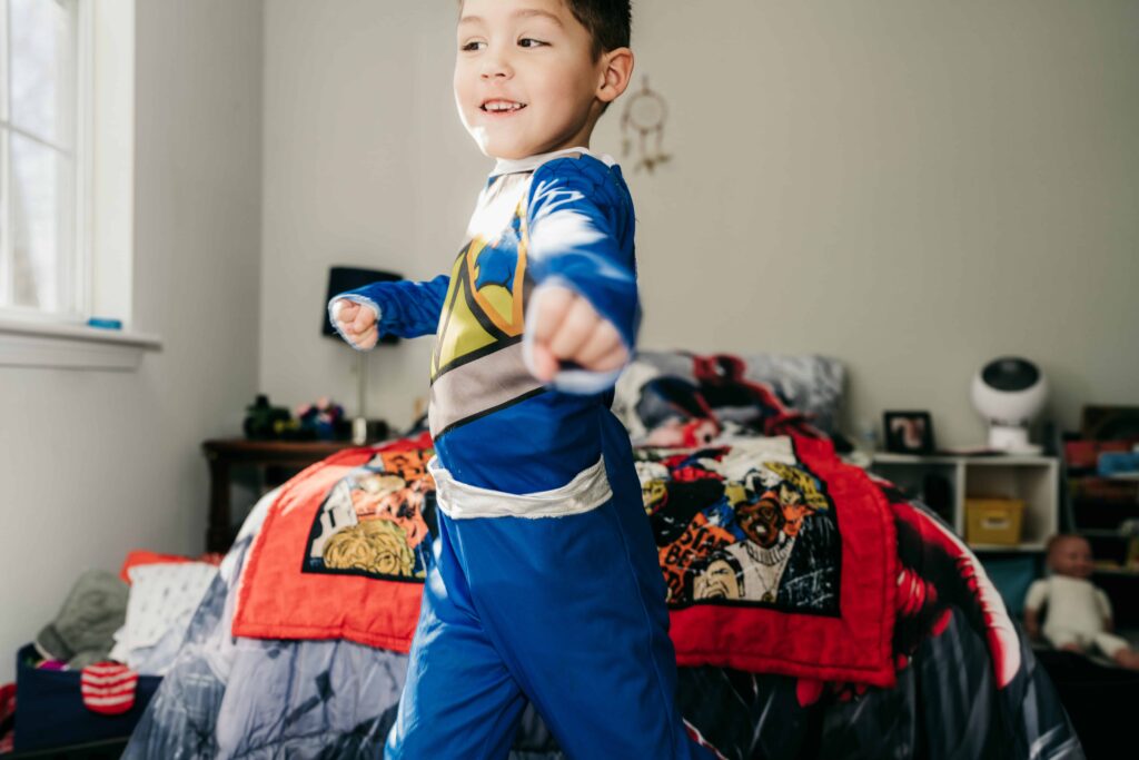 A boy dressed in a super hero costume performs a karate chop in his bedroom during a documentary family photography session by These Days Photography in Fairfax, VA